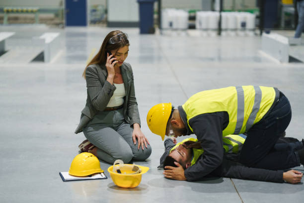 Colleagues giving first aid to unconscious worker after accident at work in warehouse, modern factory. Calling the ambulance. Concept of occupational safety and health in workplace.